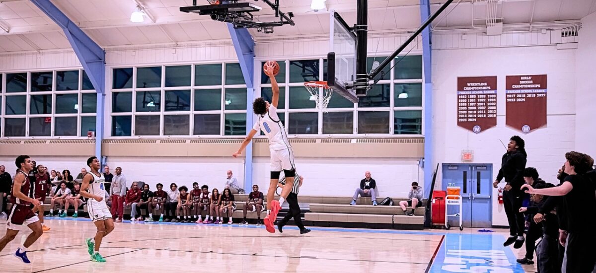 In Westtown School gym, a player in a white uniform is airborne beneath the basket, reaching up with the ball for a dunk. Teammates and oppnonents stand on the court, both team benches and spectators line the far wall, and championship banners hang on the gym wall behind them.