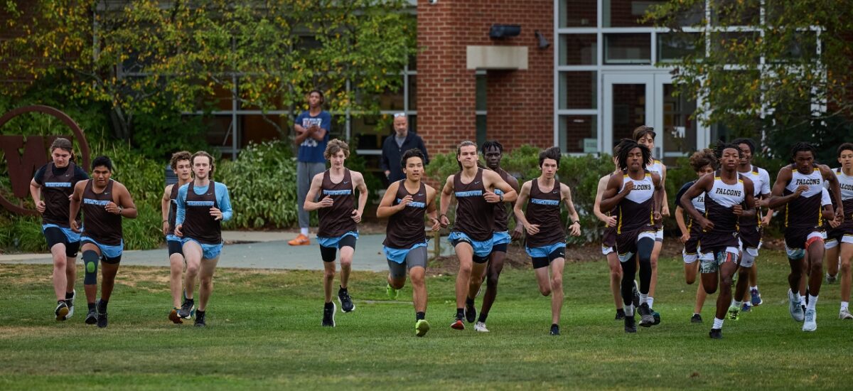 A mixed pack of boys in brown-and-blue Westtown singlets and runners in white-and-gold uniforms run across a grassy field at the start of a cross-country race, with a brick school building, shrubs and a few spectators visible in the background.