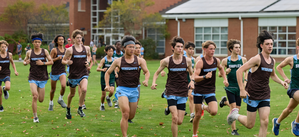 A mixed pack of boys in brown-and-blue Westtown singlets and runners in white-and-gold uniforms run across a grassy field at the start of a cross-country race, with a brick school building, shrubs and a few spectators visible in the background.