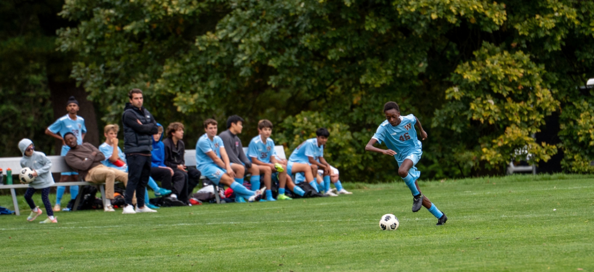 A male soccr player dribbling the ball up the sideline in a blue Westtown Soccer uniform while his teammates on the beach watch him from the sidelines.
