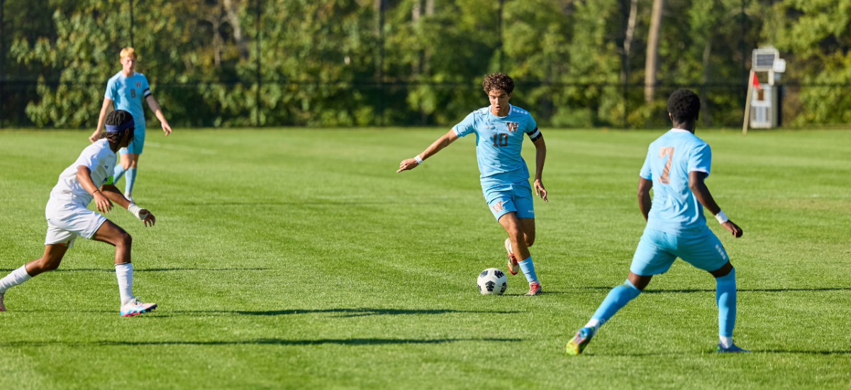 A boy in a light-blue soccer kit (number 26) dribbles a red-and-white ball across a green field while three opponents in gray jerseys move to challenge him; other players and trees line the background