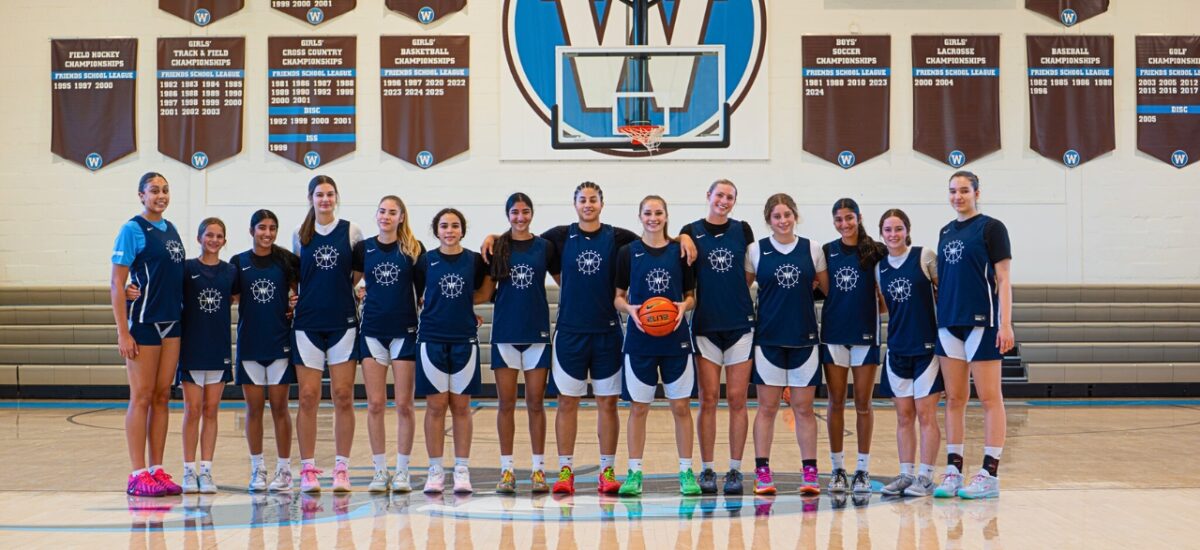Girls basketball team members posing for a team photo on a basketball court with the Westtown circle W logo and a basketball hoop in the background