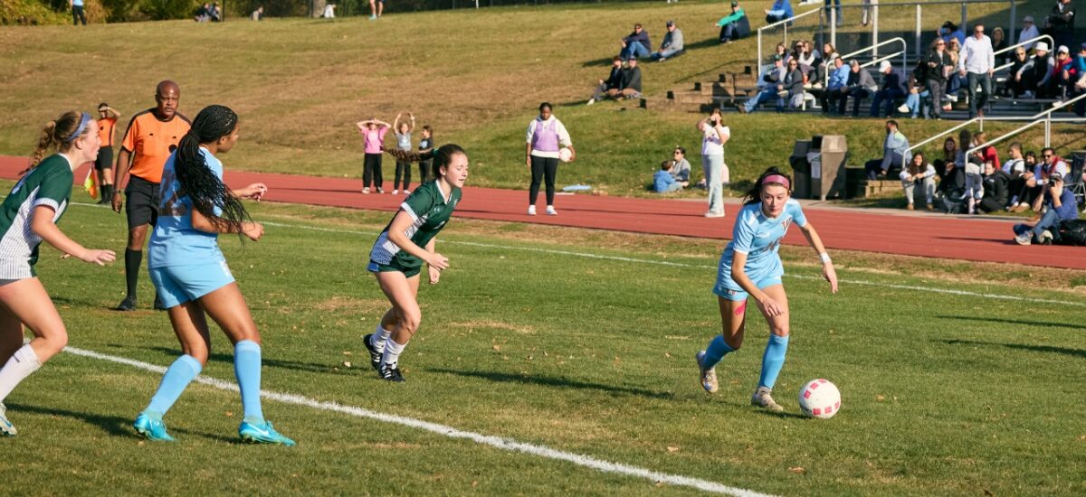 A girl in a light-blue soccer uniform dribbles a pink-and-white ball near the sideline as three opponents in dark-green kits sprint toward her; a referee, track, and spectators on bleachers watch.