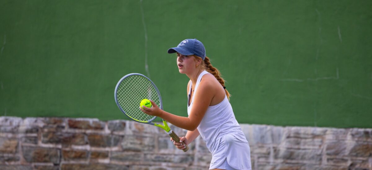 A teenage girl in a white tennis outfit and navy cap holds a yellow tennis ball in one hand and a racket in the other, eyes focused as she readies a serve in front of a green backboard and low stone wall.