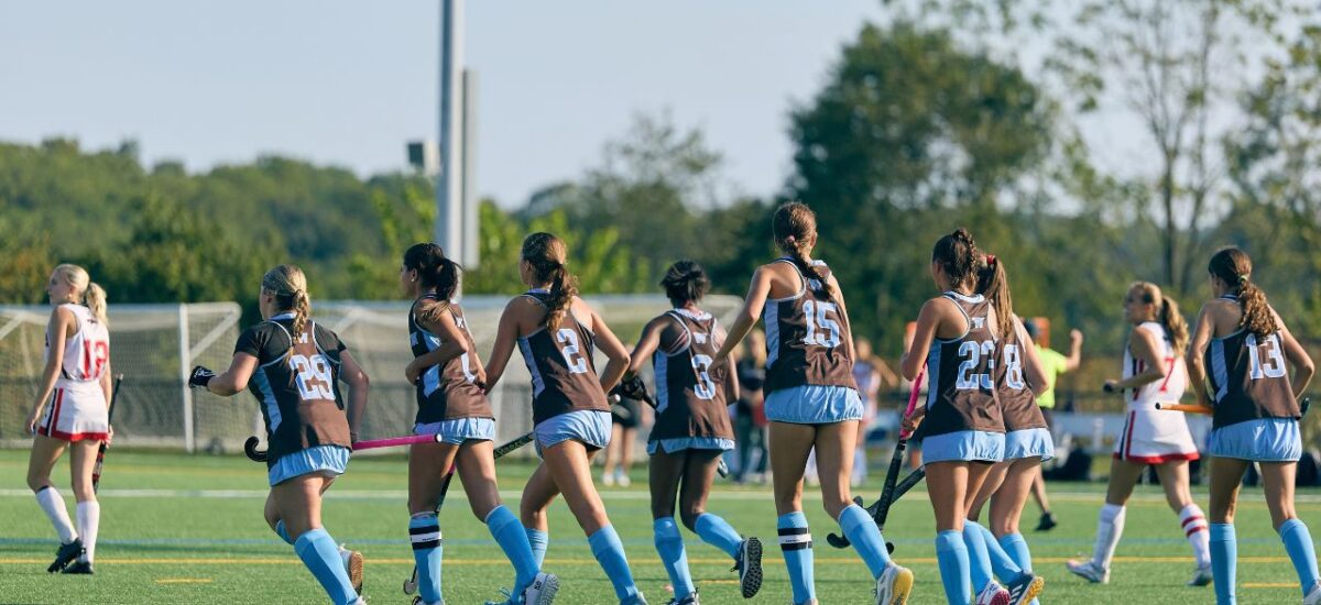 A group of teenage girls in dark brown and blue field hockey skirts jog across a turf field.