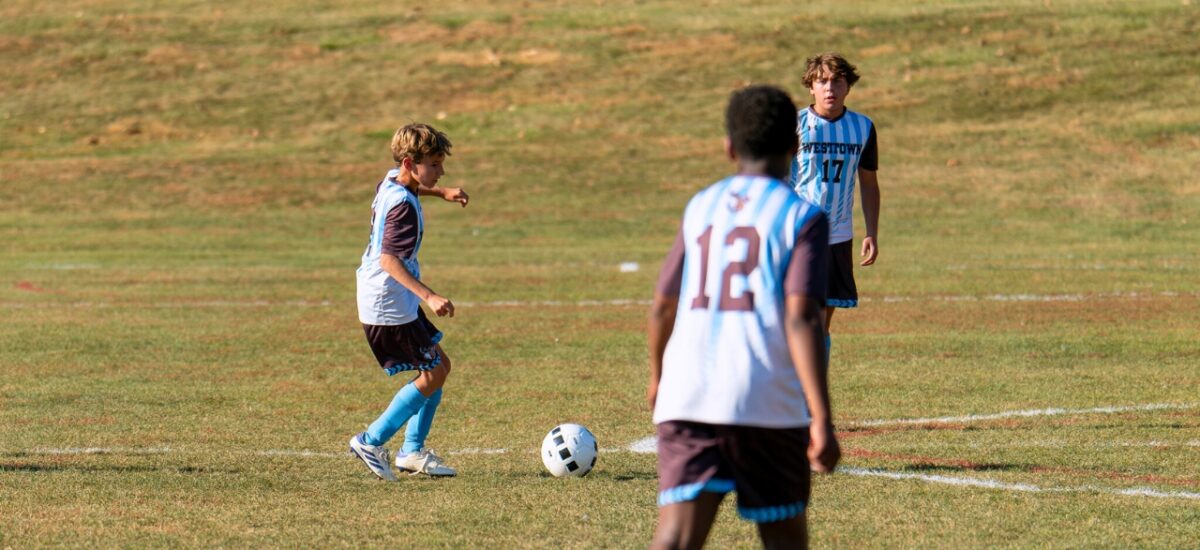 Three middle-school boys in Westtown striped jerseys on a sunlit grass field; one player dribbles the ball and readies a pass while two teammates watch and move into position.