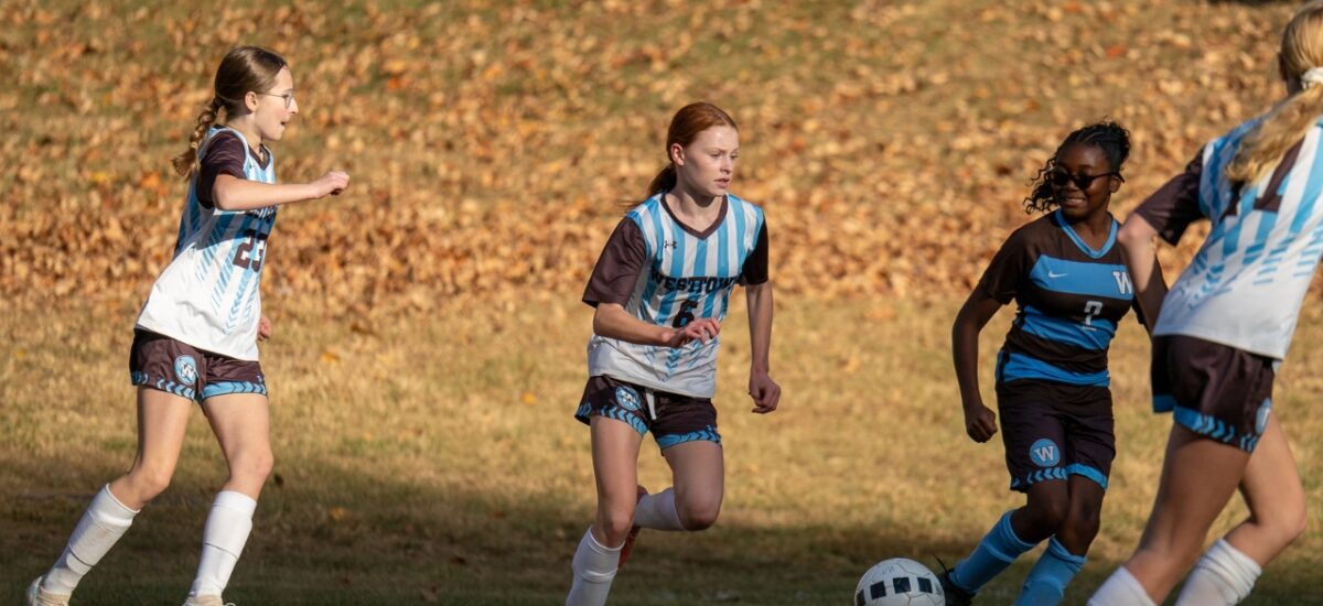 Westtown middle-school girls soccer players converge on the ball in a fall match, two players in light-blue Westtown jerseys and one opponent in dark kit contesting possession on a grass field.