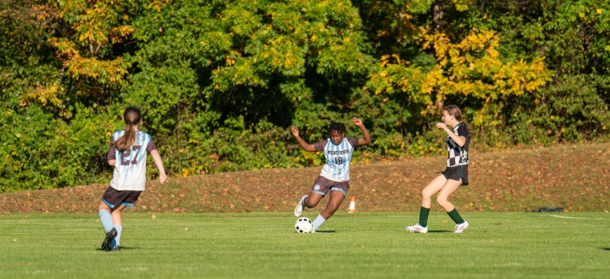 Westtown middle-school girls soccer players converge on the ball in a fall match, two players in light-blue Westtown jerseys and one opponent in dark kit contesting possession on a grass field.