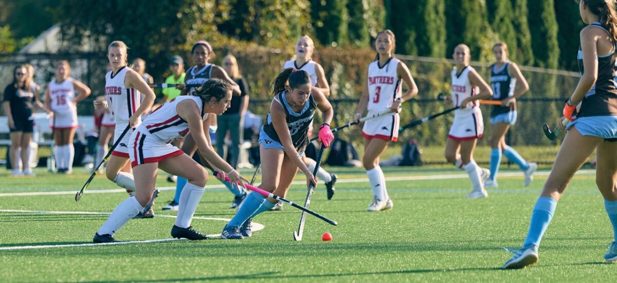 A Westtown Field Hockey student-athlete in dark brown and blue skirt Westtown uniform controls the ball while surrounded by opposing players in white uniforms on a Field Hockey Turf Field.