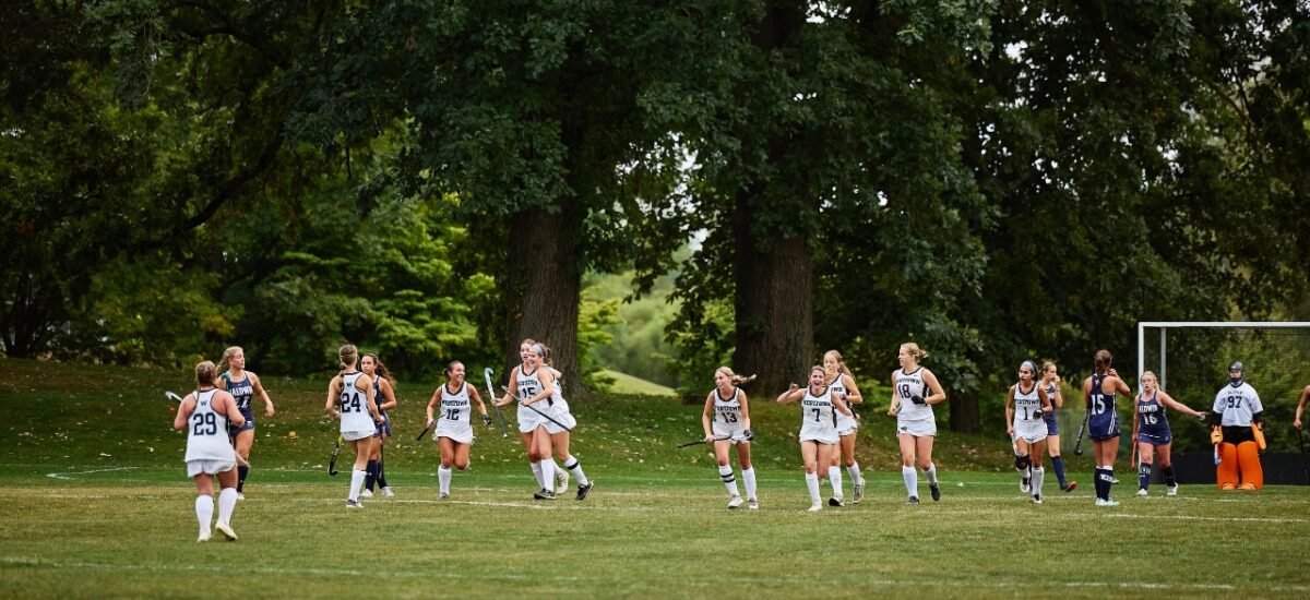 A line of girls in white Westtown field-hockey uniforms jog and celebrate across a grassy field; opponents in dark uniforms and an orange-padded goalkeeper stand by the goal with tall trees and greenery in the background.