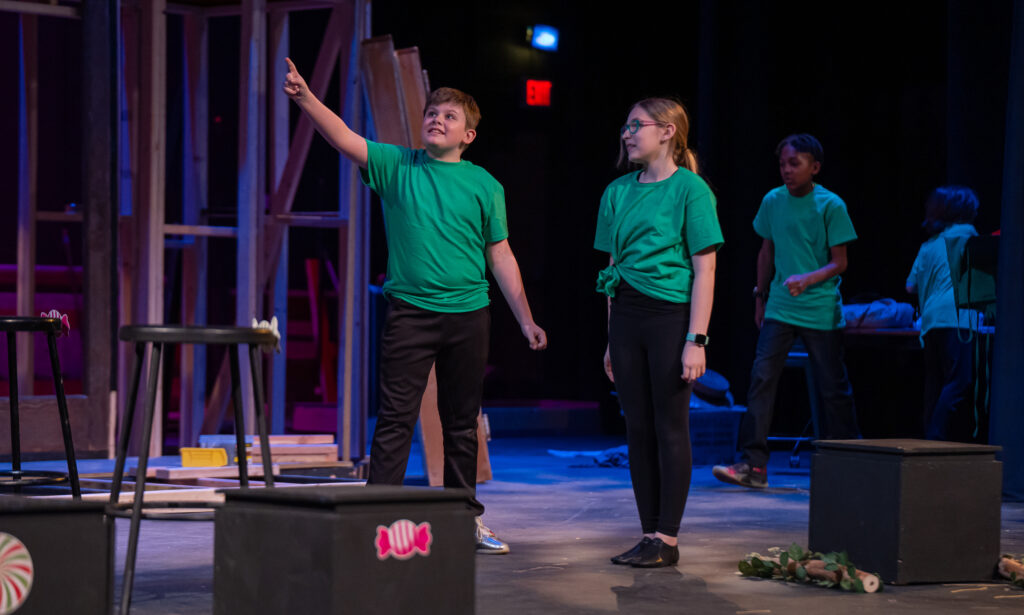 **5th Grade Play**
Westtown School 5th grade students in matching green T-shirts performing on stage amid set pieces, with one student pointing dramatically upward and classmates watching under warm theater lighting.