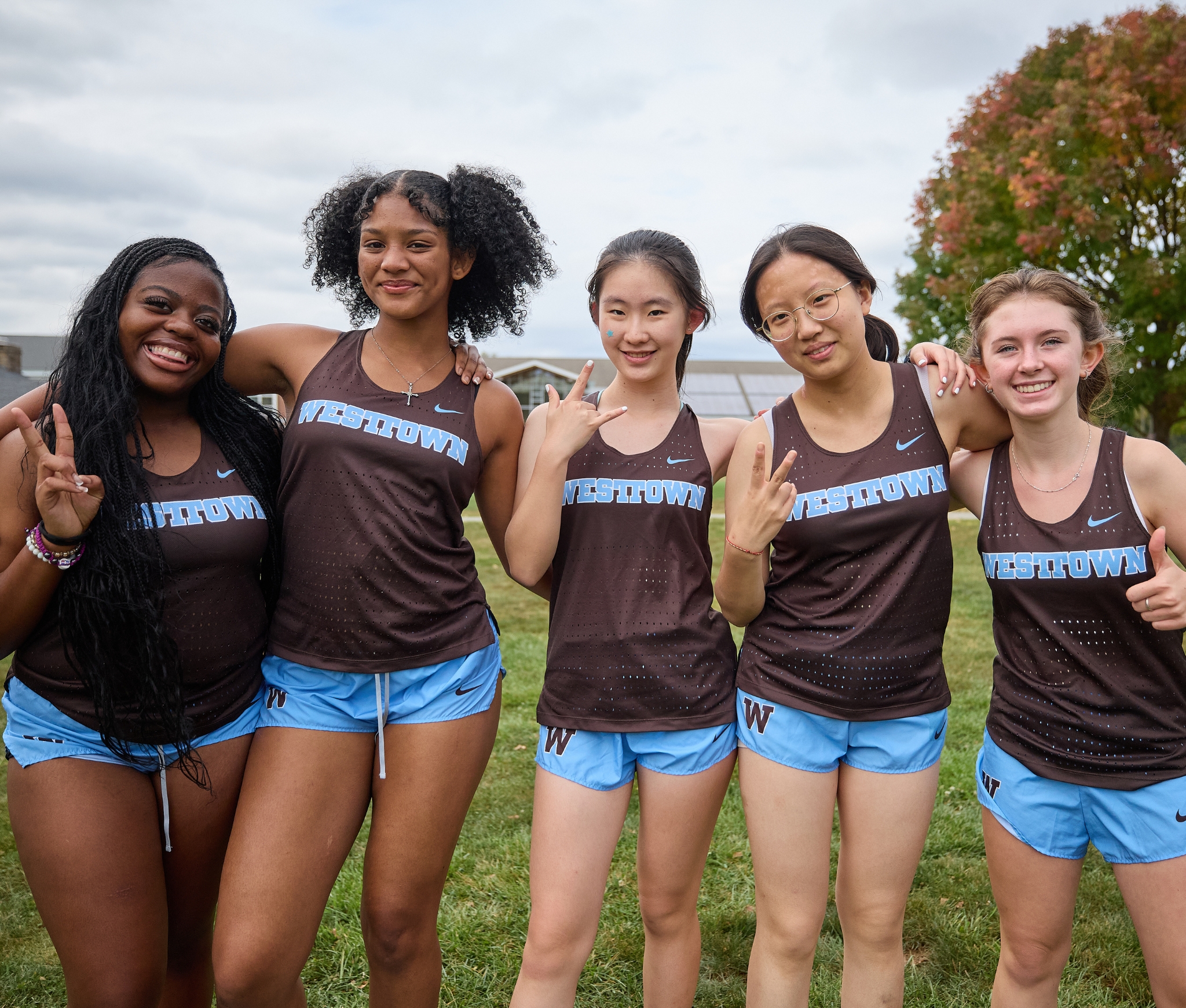 Westtown School girls cross country runners in brown-and-blue “Westtown” singlets smiling and posing together on a grassy field with autumn trees in the background