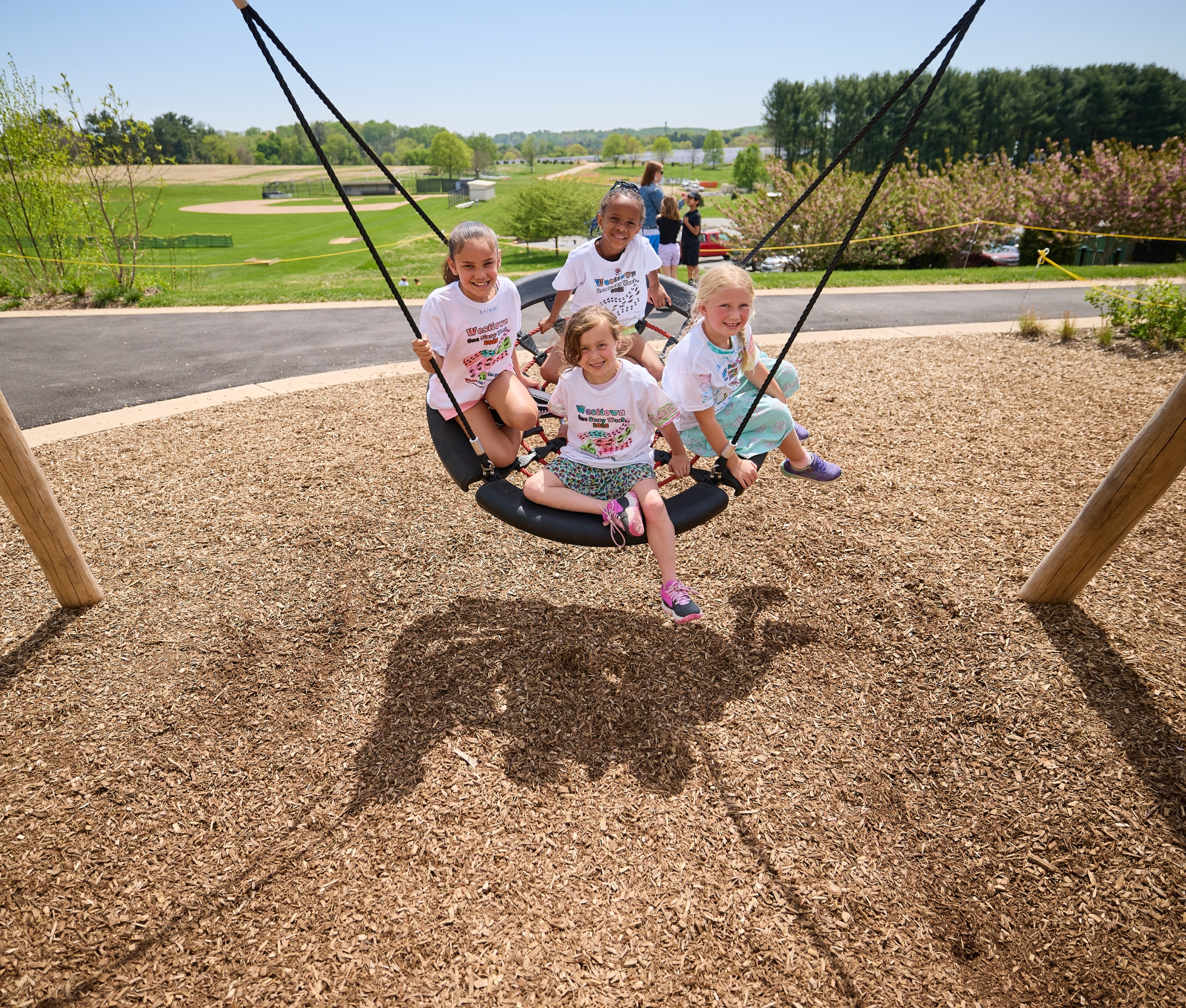 Westtown School Lower School students laughing and swinging together on a large circular playground swing with athletic fields and spring greenery in the background