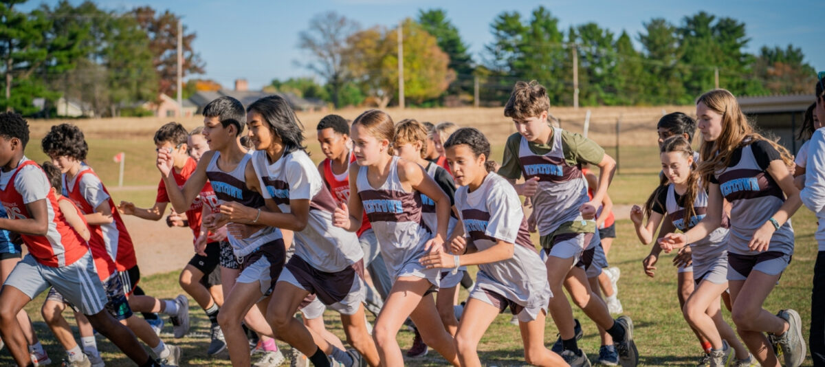Coed Westtown School middle-school cross-country runners in brown-and-blue singlets launching from the starting line at a regional cross-country meet on a grassy course — youth athletics, team spirit, and outdoor fitness.