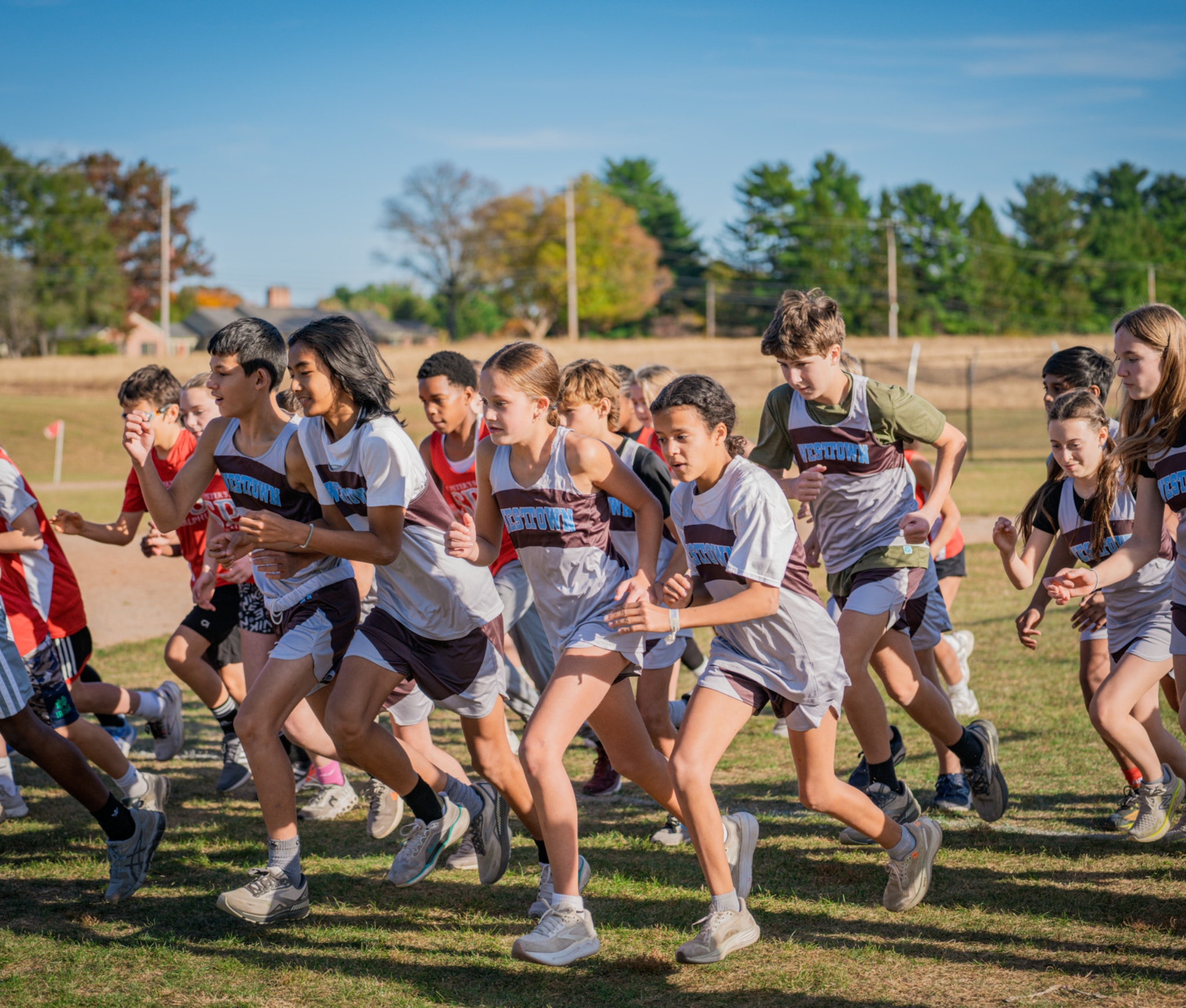 Westtown School middle school cross country runners in brown-and-white singlets sprinting from the start line across an open grassy course under a clear blue sky