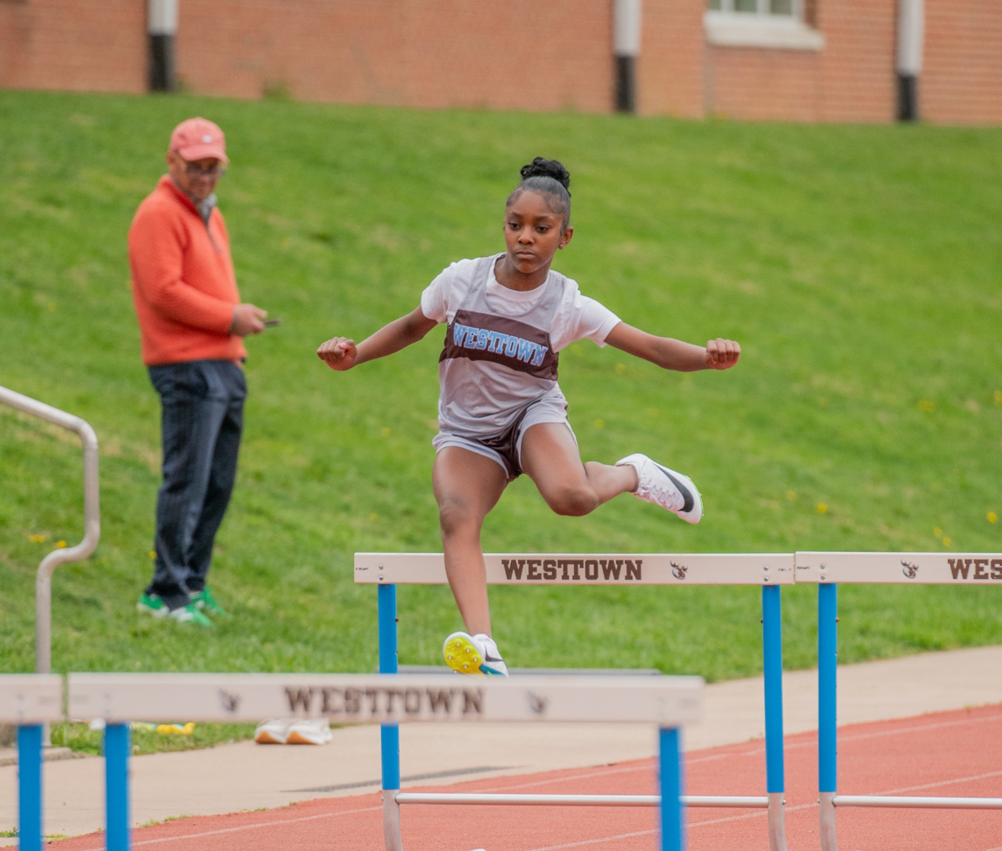 Westtown School middle-school girl hurdler in brown-and-blue singlet mid-air over a Westtown-branded hurdle during a spring track meet.