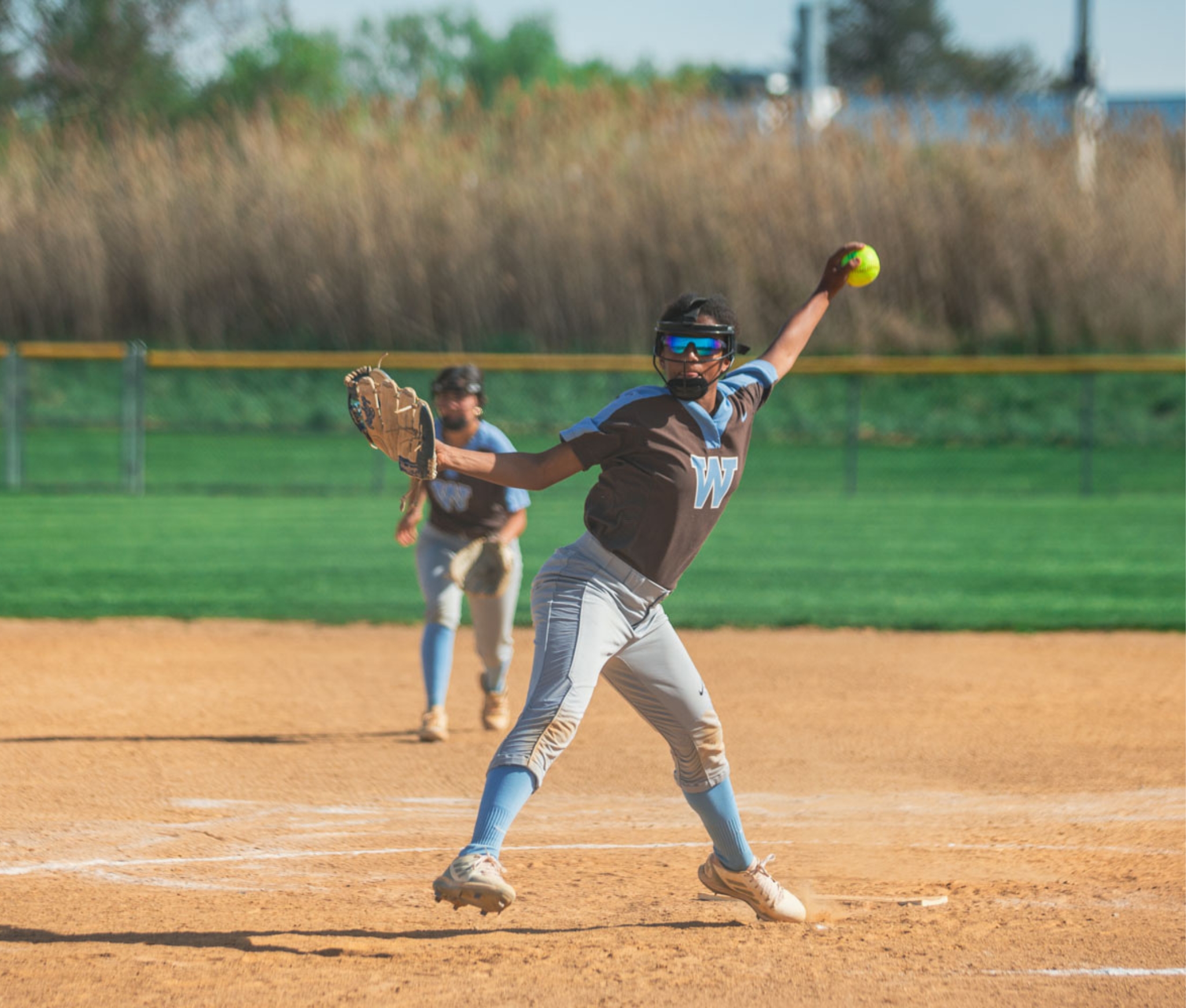 Westtown School varsity softball pitcher in brown-and-blue uniform winding up for a pitch on the diamond under late-spring sunlight