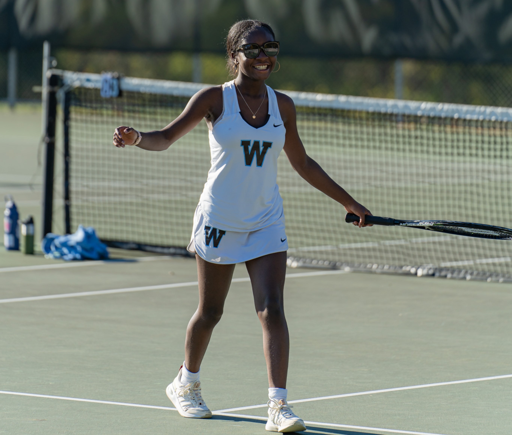 Westtown School varsity girls tennis player in white Nike dress with “W” logo smiling and holding her racket on an outdoor court with the net and wooded backdrop behind her