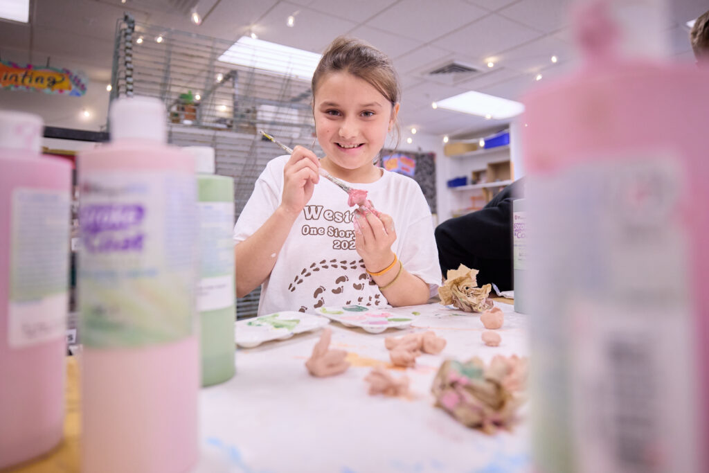 **Lower School Clay Painting**
Westtown School Lower School student smiling as she paints a hand-molded clay sculpture pink at a table in the bright art studio, surrounded by paint bottles and supplies.